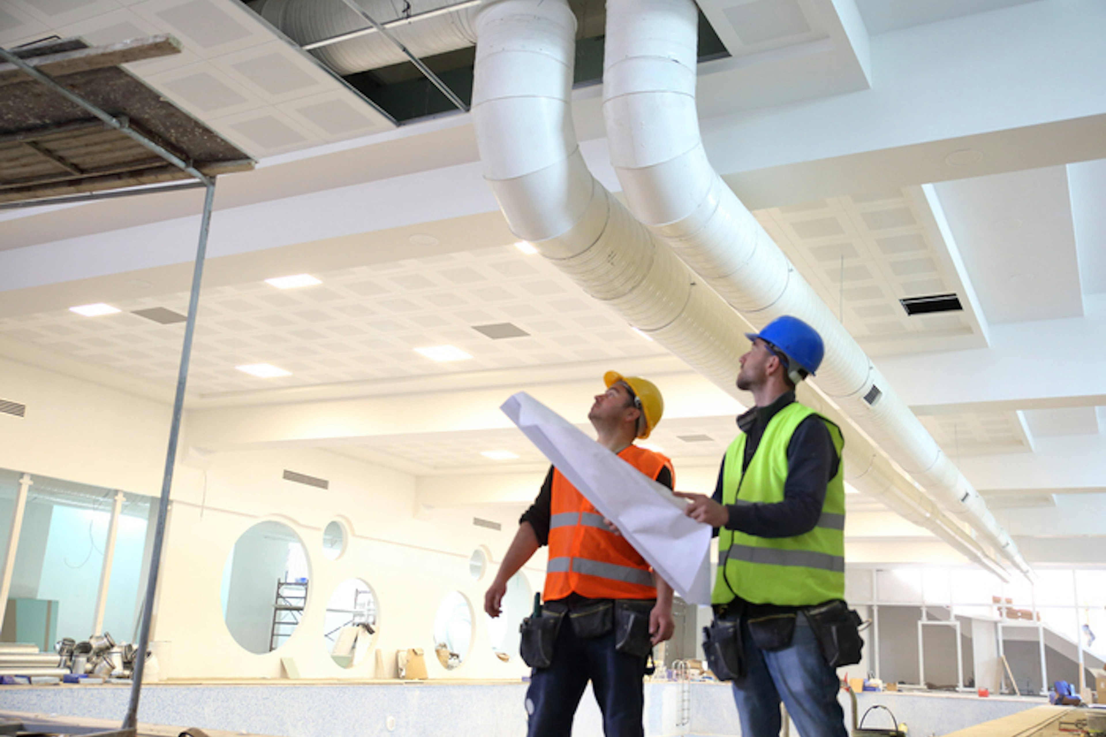 Två män i arbetskläder tittar upp mot taket i en lokal