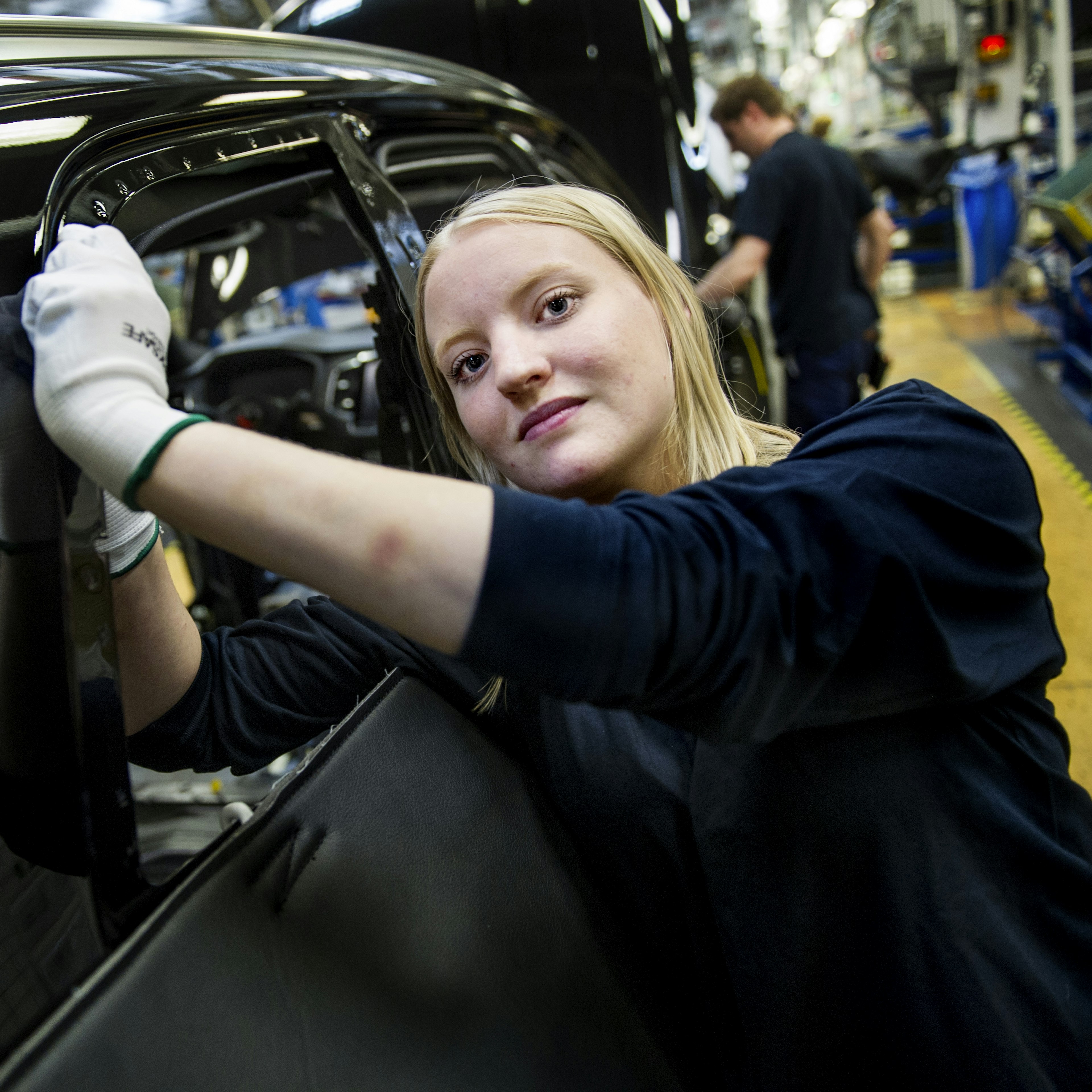 En kvinnlig arbetare tittar in i kameran samtidigt som hon arbetar med en bil i en bilfabrik