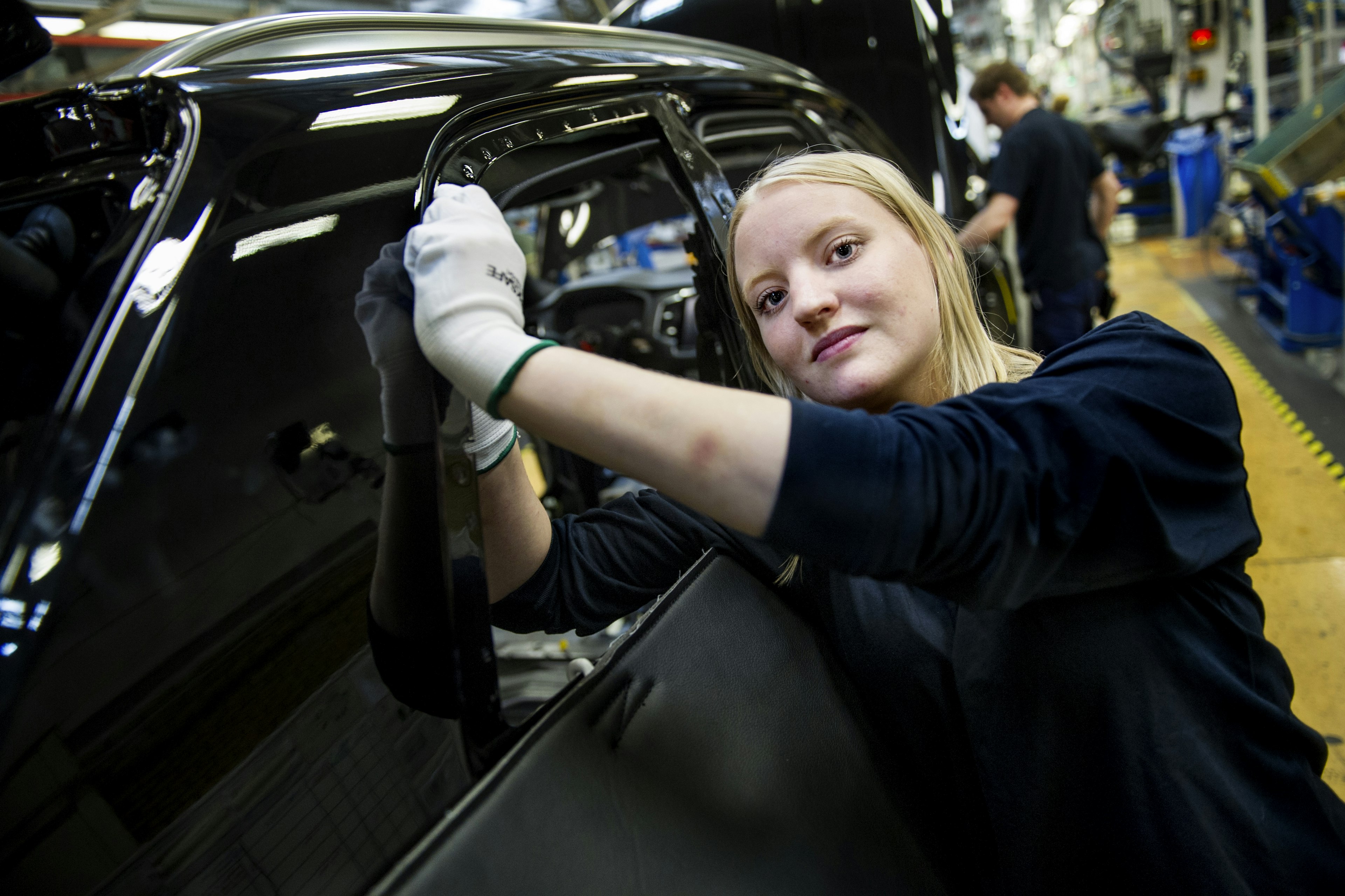 En kvinnlig arbetare tittar in i kameran samtidigt som hon arbetar med en bil i en bilfabrik
