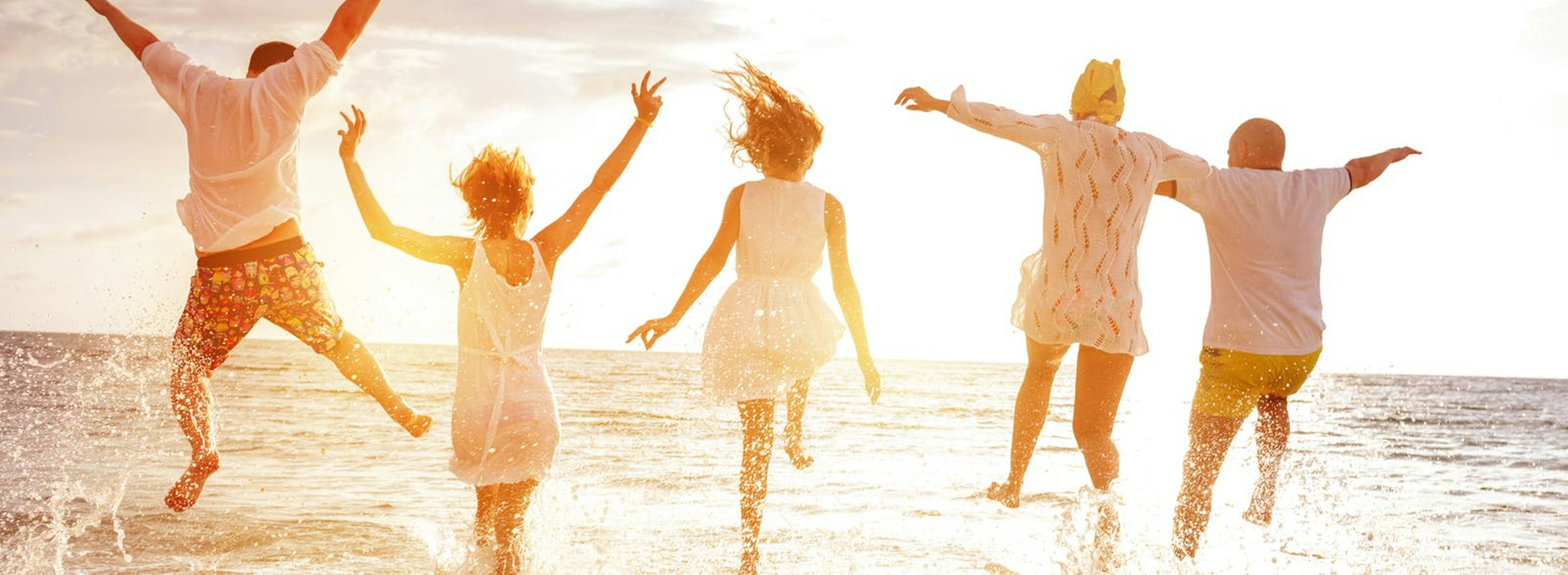 Fem personer med ryggen mot kameran som plaskar och hoppar vid stranden i solnedgången